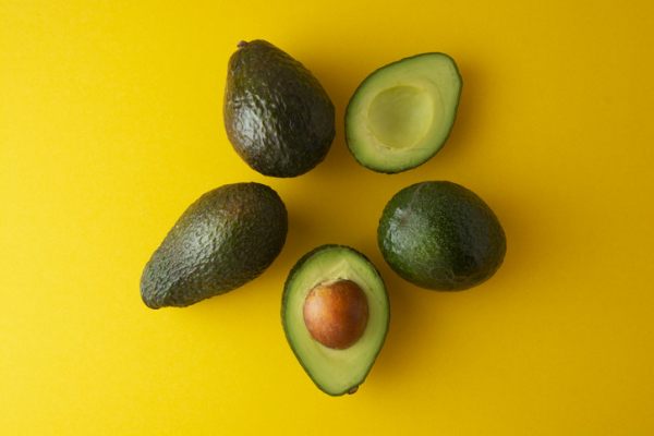 Three dark green whole avocados and two light green half avocados, one holding the pit, arranged like petals of a flower against a yellow background